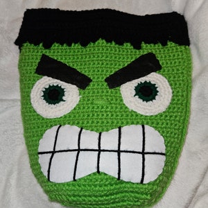 May include: Green crocheted plush toy shaped like a monster's face with black eyebrows, white eyes with green pupils, and a white mouth with black teeth.