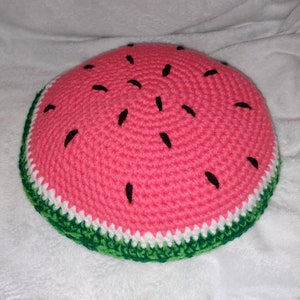 May include: A crocheted watermelon pillow with a pink center, white rind, and green outer edge. The pillow has black embroidered seeds.