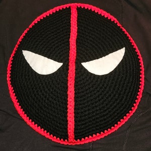 May include: A black and red crocheted pillow shaped like Deadpool's mask. The mask has white eyes and a red line down the center.