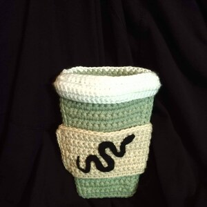 May include: A crocheted green and beige coffee cup cozy with a black snake design.