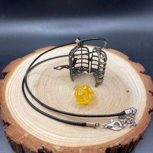 Open birdcage pendant locket necklace hung from a black cord next to a mini d20 dice displayed on a log slice