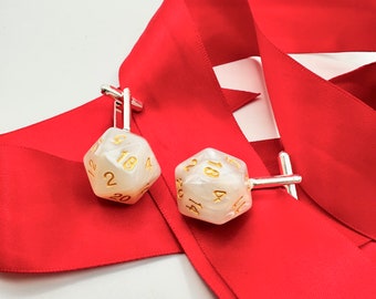 May include: A pair of cufflinks featuring white, iridescent dice with gold numbers. The dice are attached to silver cufflink backs. The cufflinks are displayed on a red ribbon.