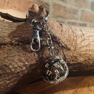 May include: A black and silver dragon keychain with a black and white dice charm. The dice has the numbers 1, 7, and 17 visible. The keychain is attached to a silver chain and a silver clip.