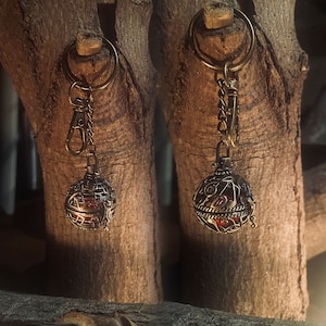 May include: Two ornate metal keychains with intricate designs and a red glass orb inside. The keychains are hanging from a wooden surface.