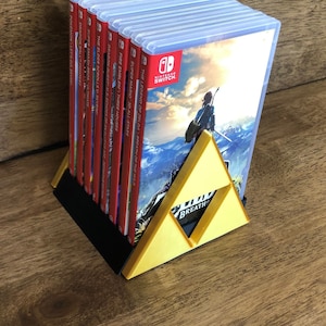 Könnte beinhalten: Ein goldener dreieckiger Ständer mit den Worten "The Legend of Zelda: Breath of the Wild" darauf gedruckt. Der Ständer hält einen Stapel Nintendo Switch-Spielkassetten, darunter Titel wie "Super Mario 3D All-Stars", "Fire Emblem: Three Houses" und "The Elder Scrolls V: Skyrim".