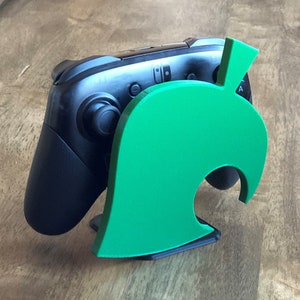 May include: A green leaf-shaped stand for a video game controller. The stand is made of plastic and has a cutout for the controller's charging port.