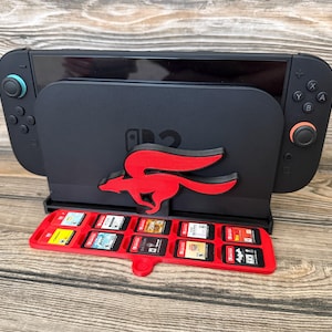May include: A black Nintendo Switch dock featuring a red fox design and a red game cartridge holder. The Switch is docked, and the holder is filled with game cartridges. The setup is on a wooden surface.