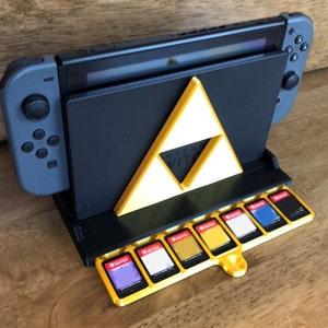 May include: A black Nintendo Switch console stand with a yellow Triforce design. The stand has a built-in storage compartment for game cartridges. The console is in a vertical position with the screen facing up. The stand is on a wooden surface.