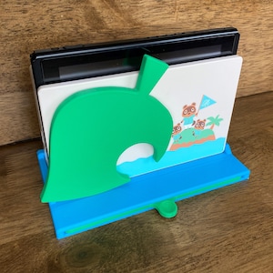 May include: A green leaf-shaped stand with a blue base holds a Nintendo Switch console. The stand features a white panel with a cartoon image of characters from the Animal Crossing video game series.