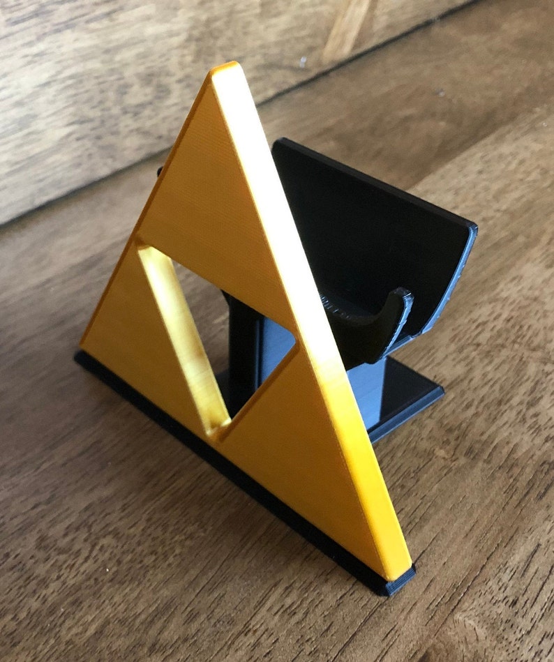 May include: A 3D printed stand for a video game console controller, shaped like the Triforce from the Legend of Zelda video game series. The stand is made of black and gold plastic.