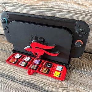 Könnte beinhalten: Eine schwarze Spielekonsolen-Dockingstation mit einem stilisierten roten Fuchsemblem auf der Vorderseite. An der Basis ist ein roter Kunststoffhalter für Spielkassetten befestigt, der zehn kleine rechteckige Spielkarten mit bunten Etiketten enthält.