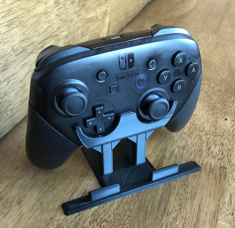 May include: A black Nintendo Switch Joy-Con controller is mounted on a black plastic stand. The controller has buttons labeled A, B, X, Y, and +.