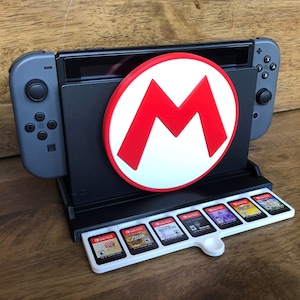 Könnte beinhalten: Eine schwarze Nintendo Switch-Konsole mit einem weißen und roten Mario-Logo auf der Vorderseite. Die Konsole steht in einem Ständer mit einem weißen und grauen Spielkartenhalter, der acht Spielkarten aufnehmen kann.