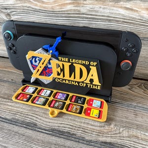 May include: A black gaming console dock featuring a yellow The Legend of Zelda Ocarina of Time logo and a blue sword and shield emblem. A yellow plastic tray attached to the front holds ten small rectangular game cartridges in individual slots.