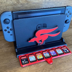 Op de afbeelding: Een zwarte Nintendo Switch-console met een rood Star Fox-logo op een zwarte standaard. De standaard heeft een sleuf voor de console en acht sleuven voor gamecartridges. De cartridges zijn rood en hebben gedrukte gametitels.