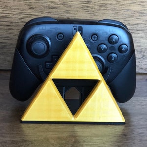 May include: A black Nintendo Switch controller sits on a yellow triangular stand. The stand is shaped like the Triforce from the Legend of Zelda video game series.