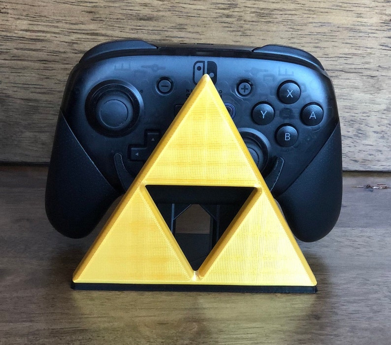 May include: A black Nintendo Switch controller sits on a yellow triangular stand. The stand is shaped like the Triforce from the Legend of Zelda video game series.