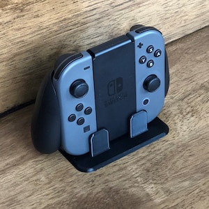 May include: A black and grey Nintendo Switch controller with a stand. The controller has a black grip and a grey face with buttons. The stand is black and has a small lip on the front to hold the controller in place.