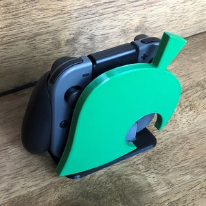 May include: A green 3D printed stand shaped like a leaf from the Animal Crossing video game. A gray Nintendo Switch controller is resting in the stand.
