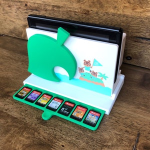 May include: White and green Nintendo Switch stand with a leaf design and a space for the console and game cartridges. The stand features a design of characters from the Animal Crossing video game.
