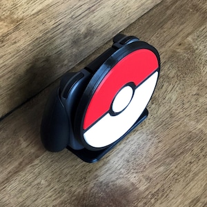 May include: Black gaming controller stand with a red, white, and black design. The stand features a circular design with a red top half and a white bottom half, with a white circle in the center. The stand is designed to hold a gaming controller.