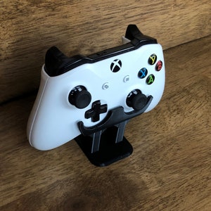 May include: A white Xbox controller is mounted on a black stand shaped like a moustache. The controller has black buttons and a black Xbox logo.