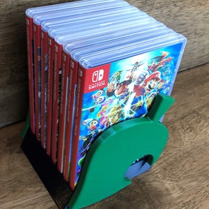 May include: A green leaf-shaped stand holds a stack of Nintendo Switch game cases. The cases are mostly red and feature titles like "Super Smash Bros. Ultimate", "Pokémon: Let's Go, Eevee!" and "Super Mario 3D All-Stars".