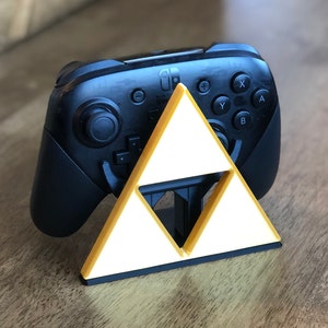 May include: A black Nintendo Switch Joy-Con controller sits in a gold Triforce stand. The stand is made of plastic and has a white interior.
