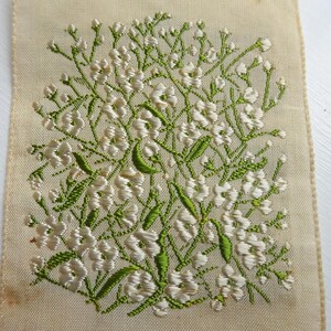 以下が含まれることがあります： 白地に緑と白の刺繍が施された花の模様のアップ。小さな白い花が緑色の茎と葉で集まっているデザインです。