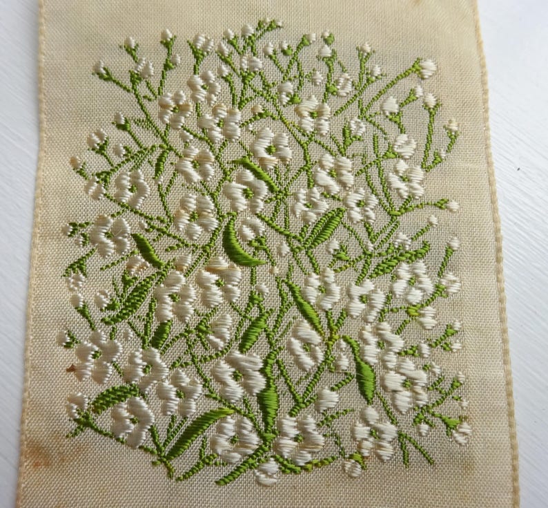 以下が含まれることがあります： 白地に緑と白の刺繍が施された花の模様のアップ。小さな白い花が緑色の茎と葉で集まっているデザインです。