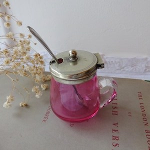 以下が含まれることがあります： 銀色の蓋と小さなスプーンが付いたピンク色のガラス製の砂糖入れ。砂糖入れは持ち手があり、本の上に置かれています。