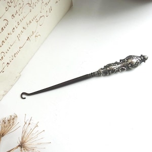 May include: An antique silver-toned button hook with an ornate handle and a dark, slender hook. The handle features detailed scrollwork and a small, decorative element. The hook is positioned near a handwritten document.