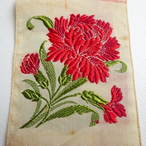 以下が含まれることがあります： 白い布地に、緑の葉と赤いカーネーションが刺繍されている