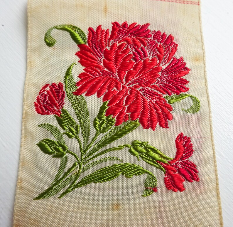 以下が含まれることがあります： 白い布地に、緑の葉と赤いカーネーションが刺繍されている