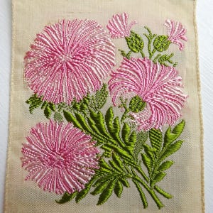 以下が含まれることがあります： ピンクと緑の花が刺繍されたヴィンテージの布パッチのクローズアップ。花はスタイリッシュで、質感があります。布はクリーム色です。
