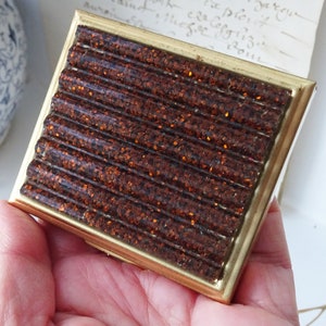 Puede incluir: Una pequeña caja rectangular de color dorado con una tapa marrón texturizada y brillante. La tapa tiene un diseño acanalado.