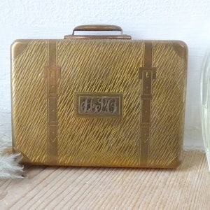 Pode incluir: Uma mala vintage de metal dourado com um padrão diagonal texturizado. A mala tem uma alça e correias decorativas. As iniciais "H.S.P.G." são exibidas em uma moldura retangular na frente.