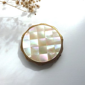 Puede incluir: Un compacto redondo de color dorado con un diseño de nácar. El compacto tiene un patrón de tablero de ajedrez y un borde ligeramente elevado.