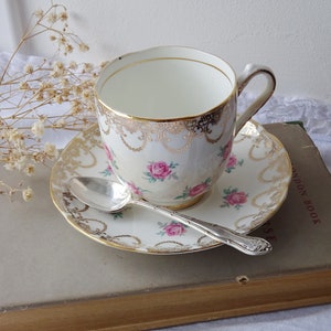 Puede incluir: Un juego de taza y platillo vintage con una cuchara de plata. El juego presenta un diseño floral con rosas rosas y ribetes dorados. La taza y el platillo están sobre un libro antiguo.