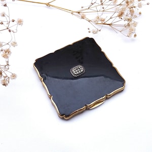 Op de afbeelding: Een zwart emaille compact met een gouden rand en een klein, zilveren marquetterie-ontwerp in het midden.