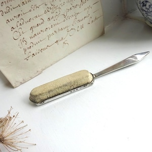 Op de afbeelding: Een zilveren briefopener met een rechthoekig, crèmekleurig kussen en een puntig mes. De opener ligt op een wit oppervlak naast een handgeschreven document met vervaagde inkt. Een gedroogde plant en een keramisch stuk zijn ook zichtbaar.