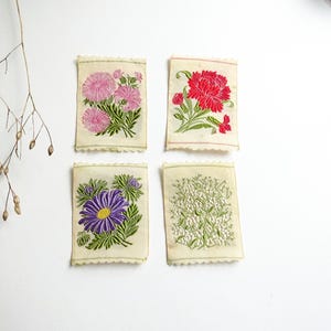 以下が含まれることがあります： 刺繍された花のデザインが施されたヴィンテージの布の正方形が4つ。正方形には、ピンクと白の花、赤い花、紫の花、白い花が描かれています。