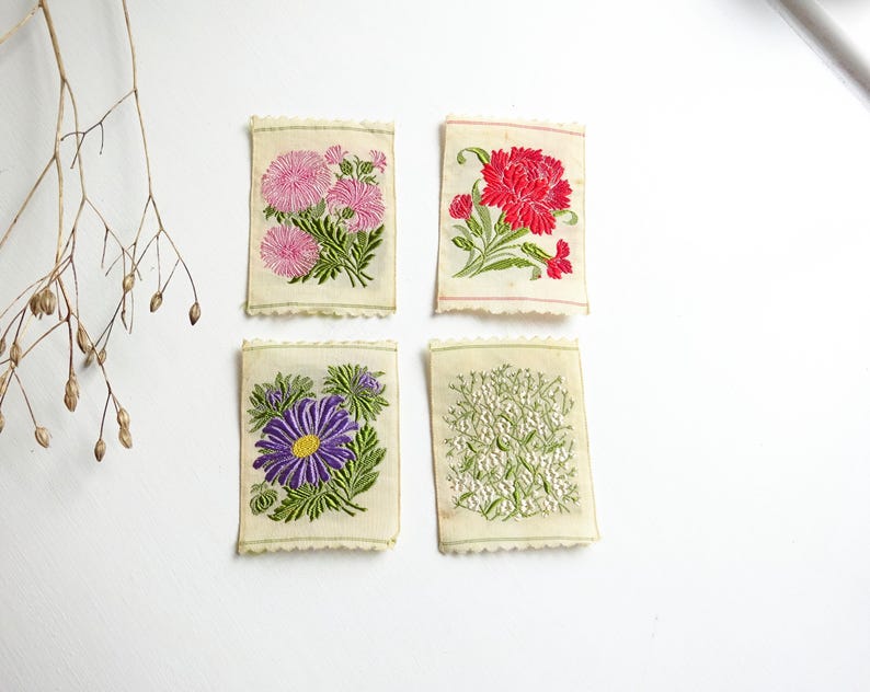 以下が含まれることがあります： 刺繍された花のデザインが施されたヴィンテージの布の正方形が4つ。正方形には、ピンクと白の花、赤い花、紫の花、白い花が描かれています。