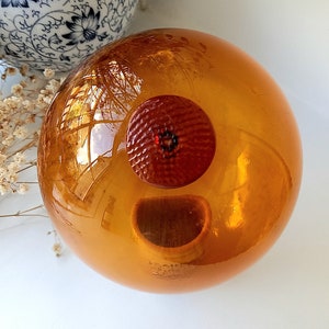 Puede incluir: Una gran esfera de vidrio de color ámbar con una pequeña abertura circular roja en el centro.