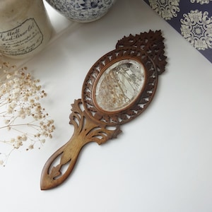 Puede incluir: Un espejo de mano de madera vintage con un marco tallado ornamentado y un pequeño espejo ovalado. El espejo está ligeramente deslustrado y tiene un aspecto rústico.