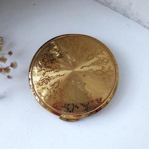 May include: A gold-toned compact with a floral design. The compact has a hinged lid and a round shape.