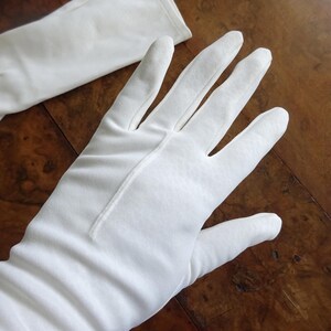 Könnte beinhalten: Ein Paar weiße Handschuhe, eine Hand geöffnet, die andere flach liegend. Die Handschuhe sind aus einem glatten, undurchsichtigen Stoff gefertigt. Die Handschuhe sind lang und reichen über das Handgelenk hinaus. Der Hintergrund ist eine dunkle Holzoberfläche.