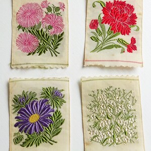 以下が含まれることがあります： 花柄の刺繍が施された布の正方形が4つあります。最初の正方形には、緑の葉が付いたピンクの花が描かれています。2番目の正方形には、緑の葉が付いた赤い花が描かれています。3番目の正方形には、緑の葉が付いた紫の花が描かれています。4番目の正方形には、緑の葉が付いた白い花が描かれています。
