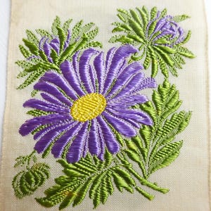 以下が含まれることがあります： クリーム色の生地に、紫と黄色の刺繍で花柄が施されています。花は緑の葉で囲まれています。