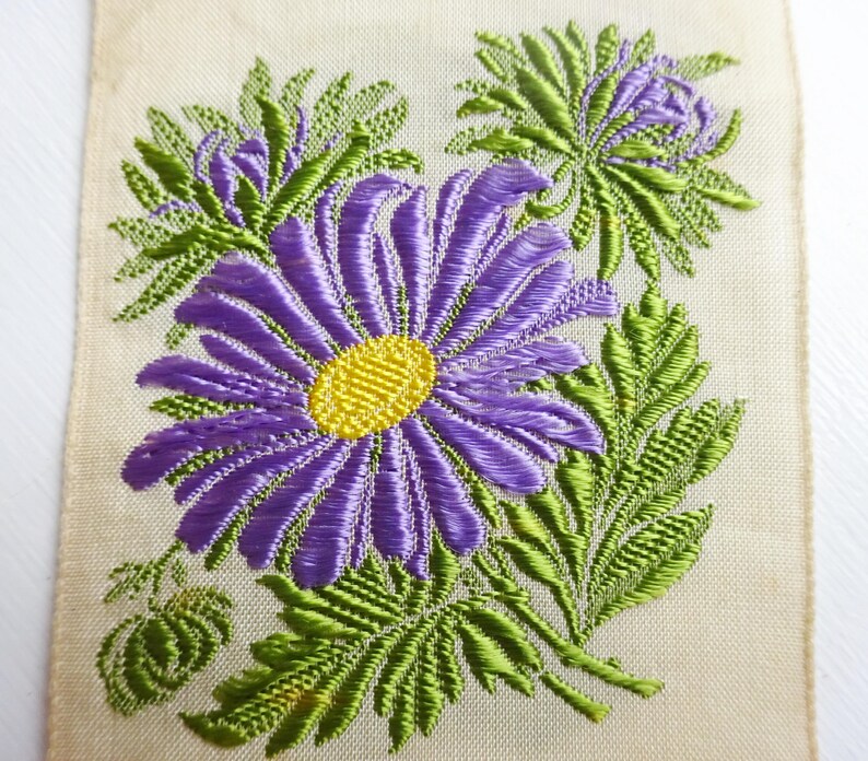 以下が含まれることがあります： クリーム色の生地に、紫と黄色の刺繍で花柄が施されています。花は緑の葉で囲まれています。